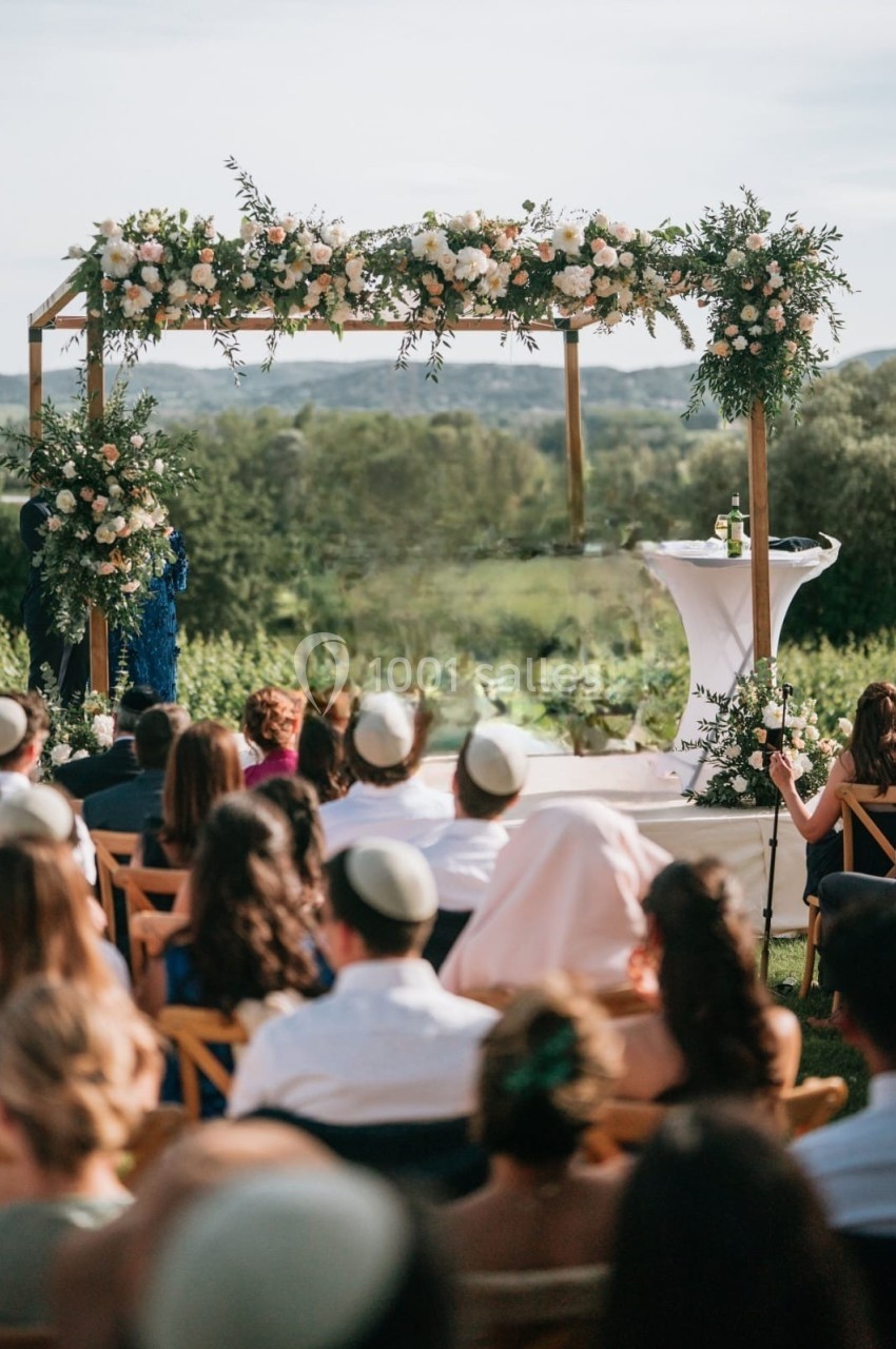 Cérémonie de mariage en plein air sous une houppa décorée de fleurs, avec des invités assis face au paysage.