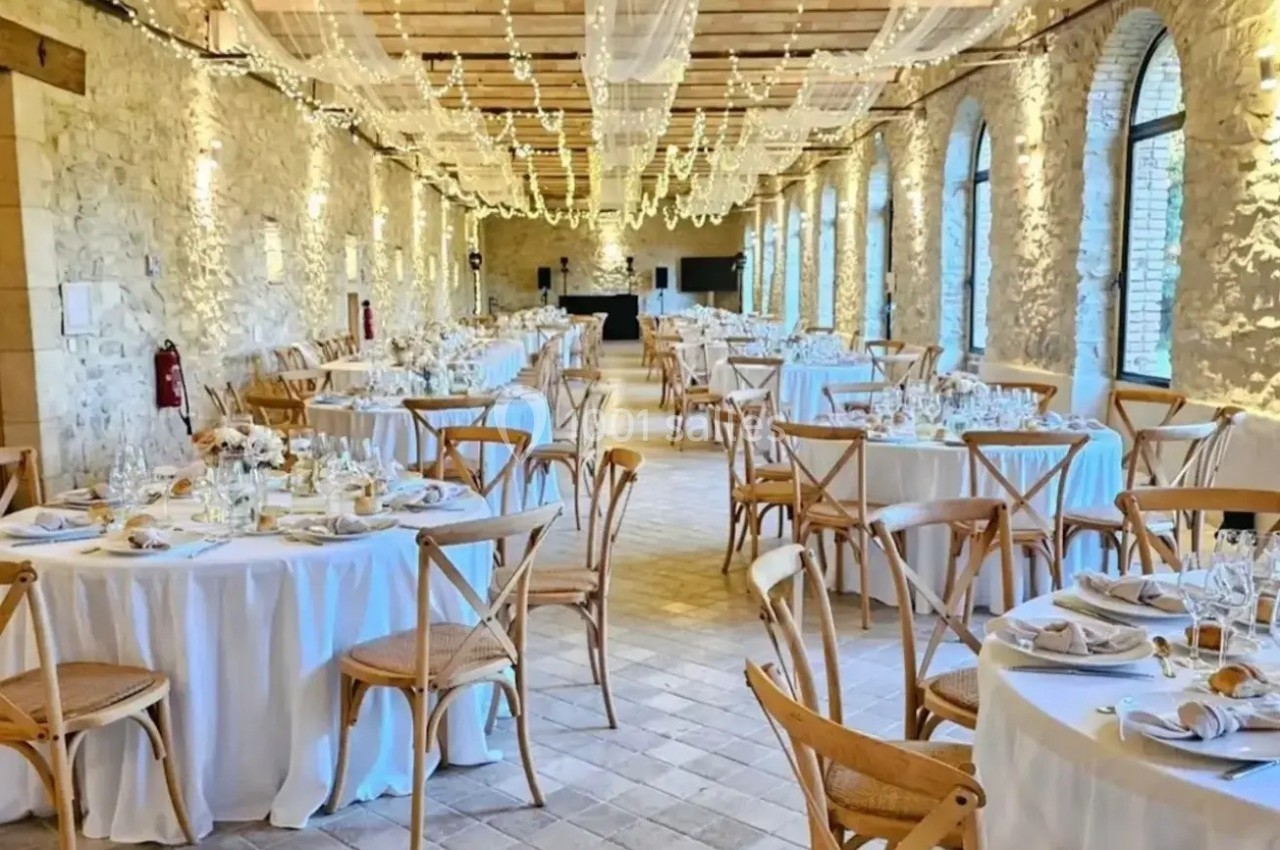 Salle de réception en pierre avec tables rondes dressées, chaises en bois et guirlandes lumineuses suspendues.
