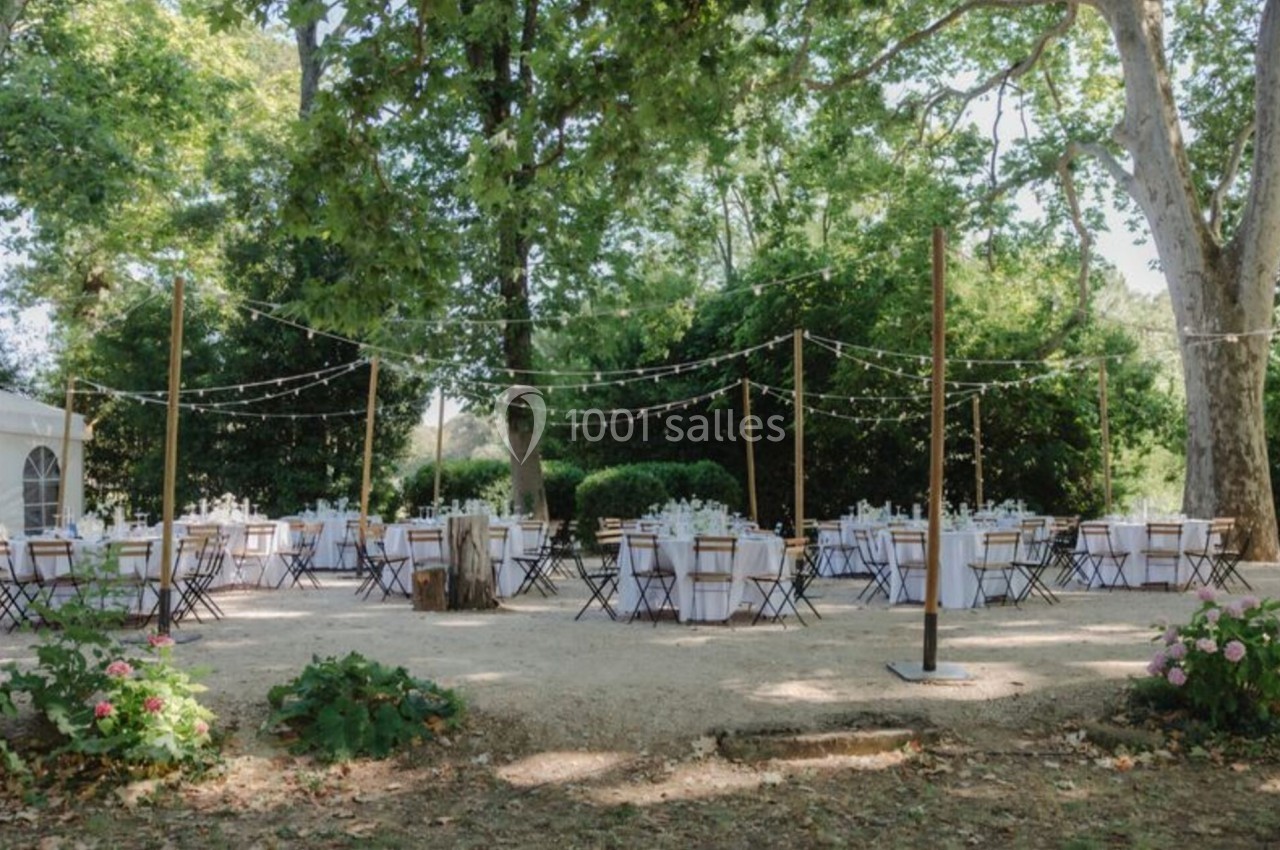Tables rondes décorées pour un événement en extérieur, sous des guirlandes lumineuses, entourées de verdure.