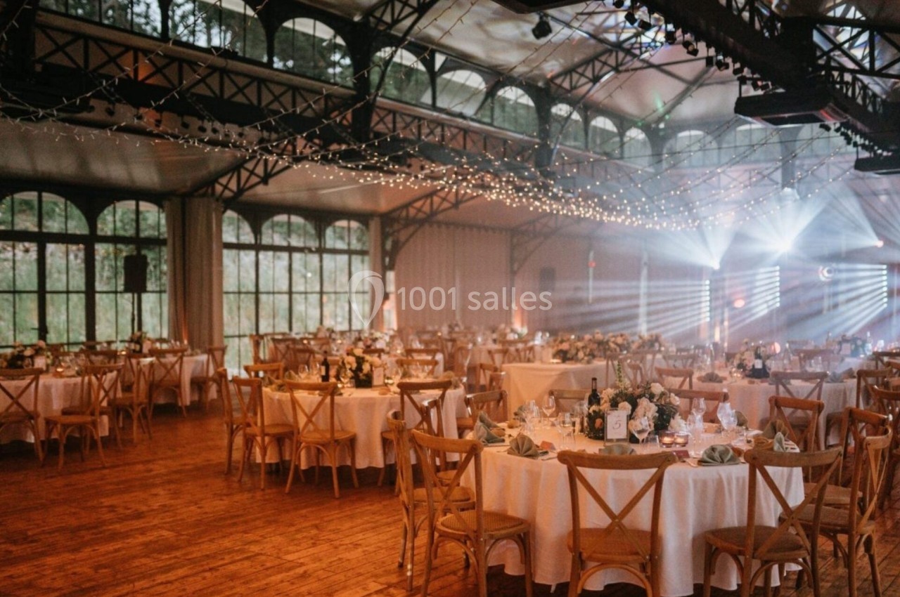 Salle de réception décorée avec des guirlandes lumineuses, tables rondes dressées et grandes fenêtres en arrière-plan.
