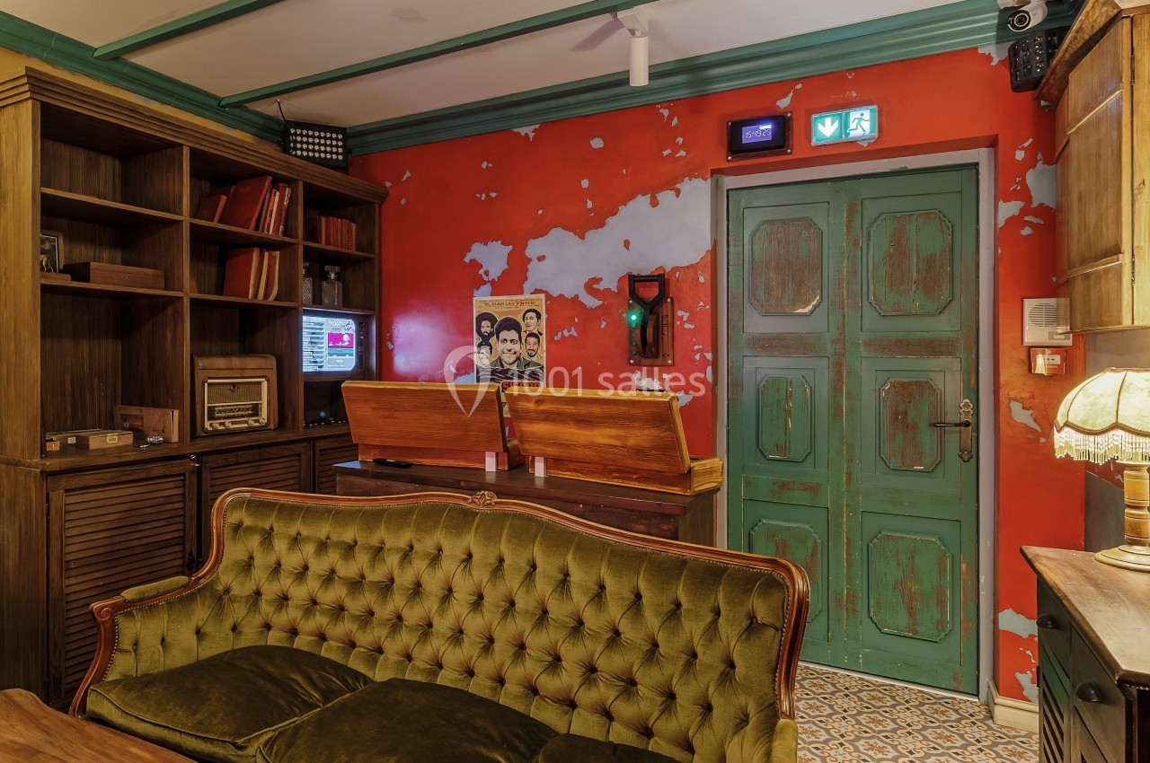 Salon vintage avec canapé vert, meubles en bois, mur rouge usé, porte verte et objets rétro comme une radio et un téléphone.
