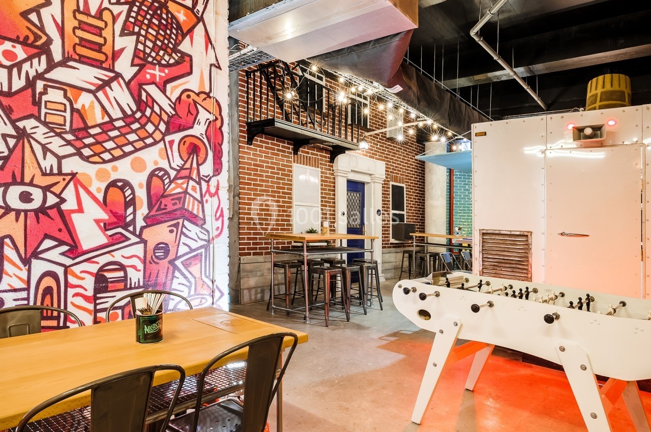 Salle avec décoration urbaine, tables en bois, baby-foot blanc et mur coloré avec graffitis.