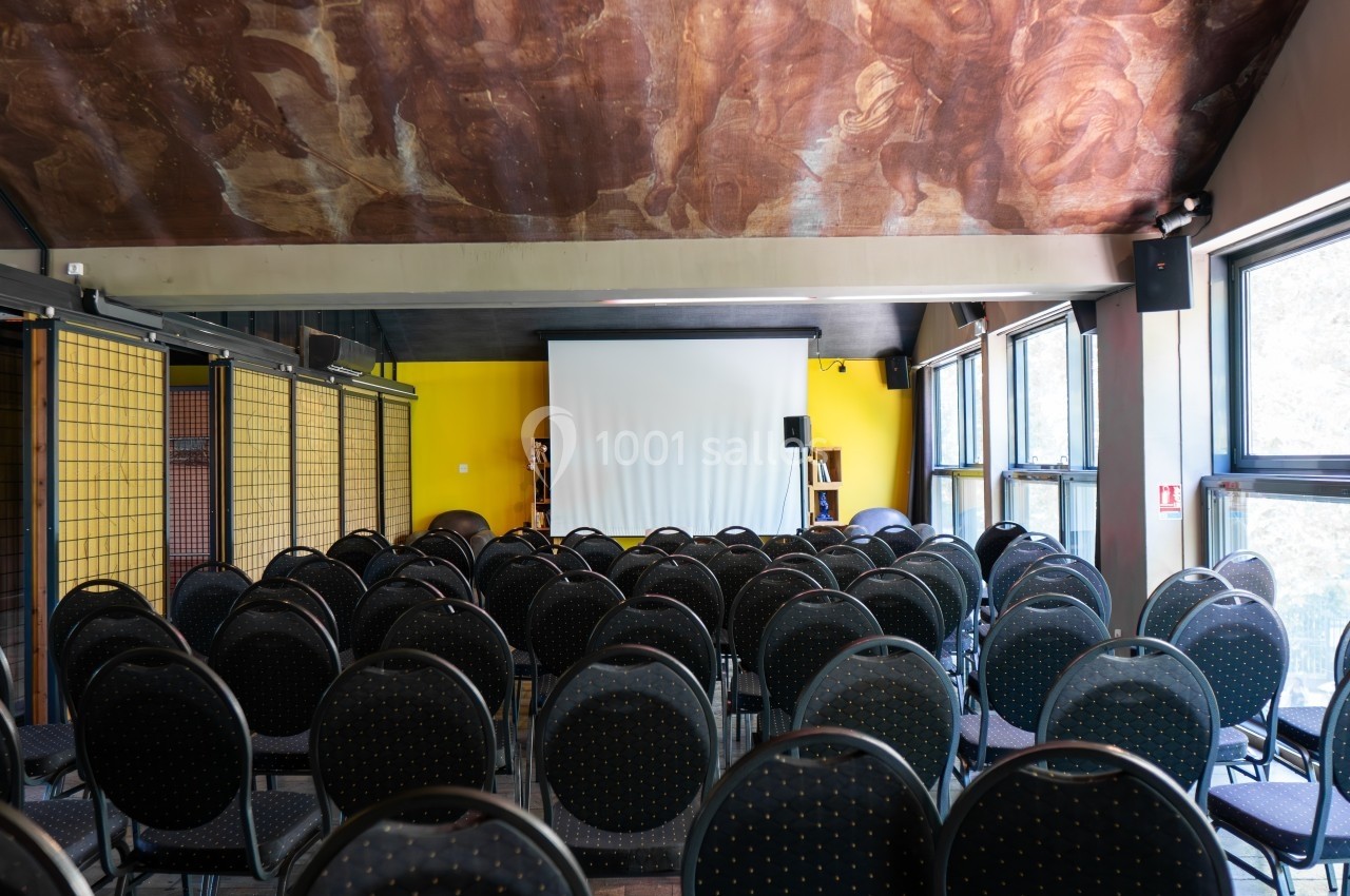 Salle de conférence vide avec des rangées de chaises noires face à un écran de projection blanc.