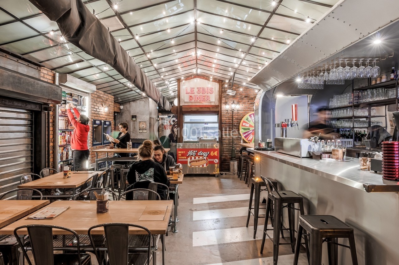 Intérieur d'un restaurant avec un bar moderne, des tables en bois, des chaises métalliques et un décor lumineux industriel.