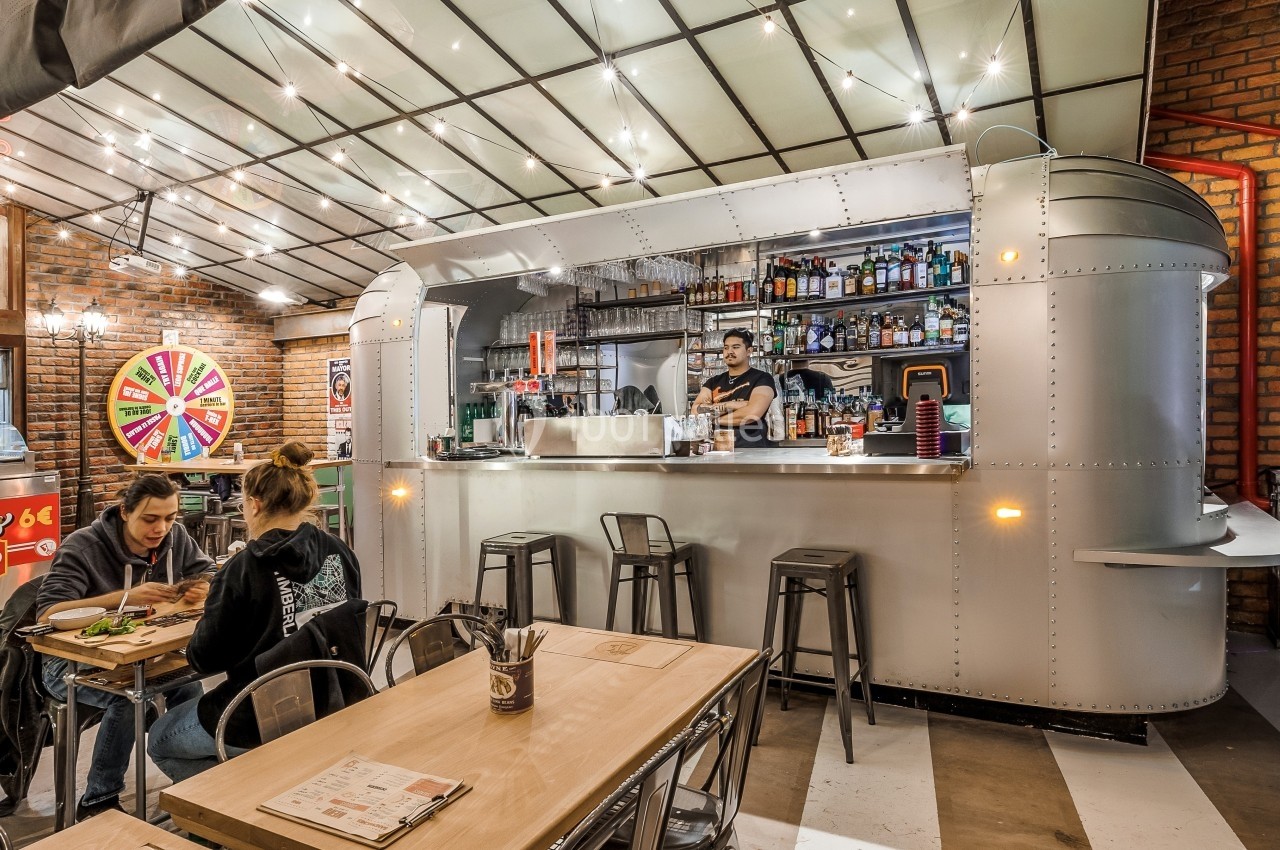 Intérieur d'un restaurant avec un bar en métal, des tables en bois, des clients attablés et un barman en service.