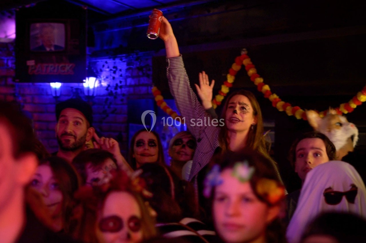 Foule déguisée lors d'une fête, une femme lève une canette dans une ambiance sombre et festive.
