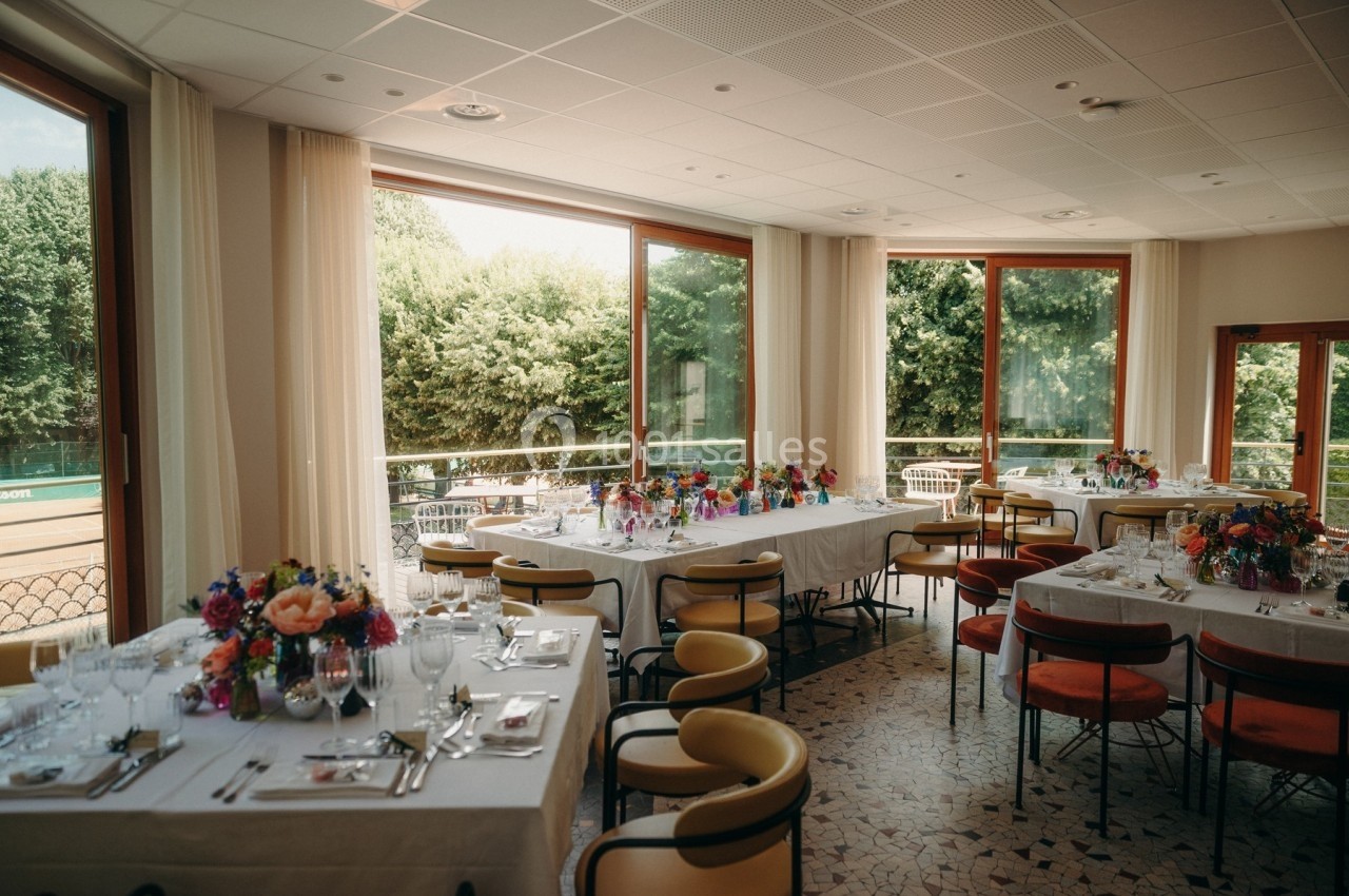 Salle lumineuse avec tables dressées pour un repas, décorées de fleurs colorées, donnant sur une terrasse et un jardin.