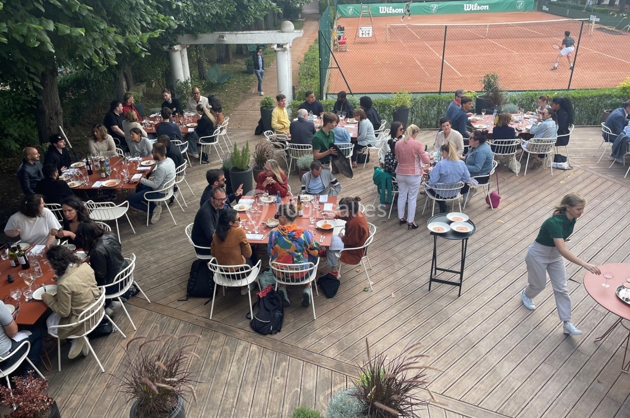 Terrasse animée avec des convives attablés, entourée de verdure, près d'un court de tennis en arrière-plan.