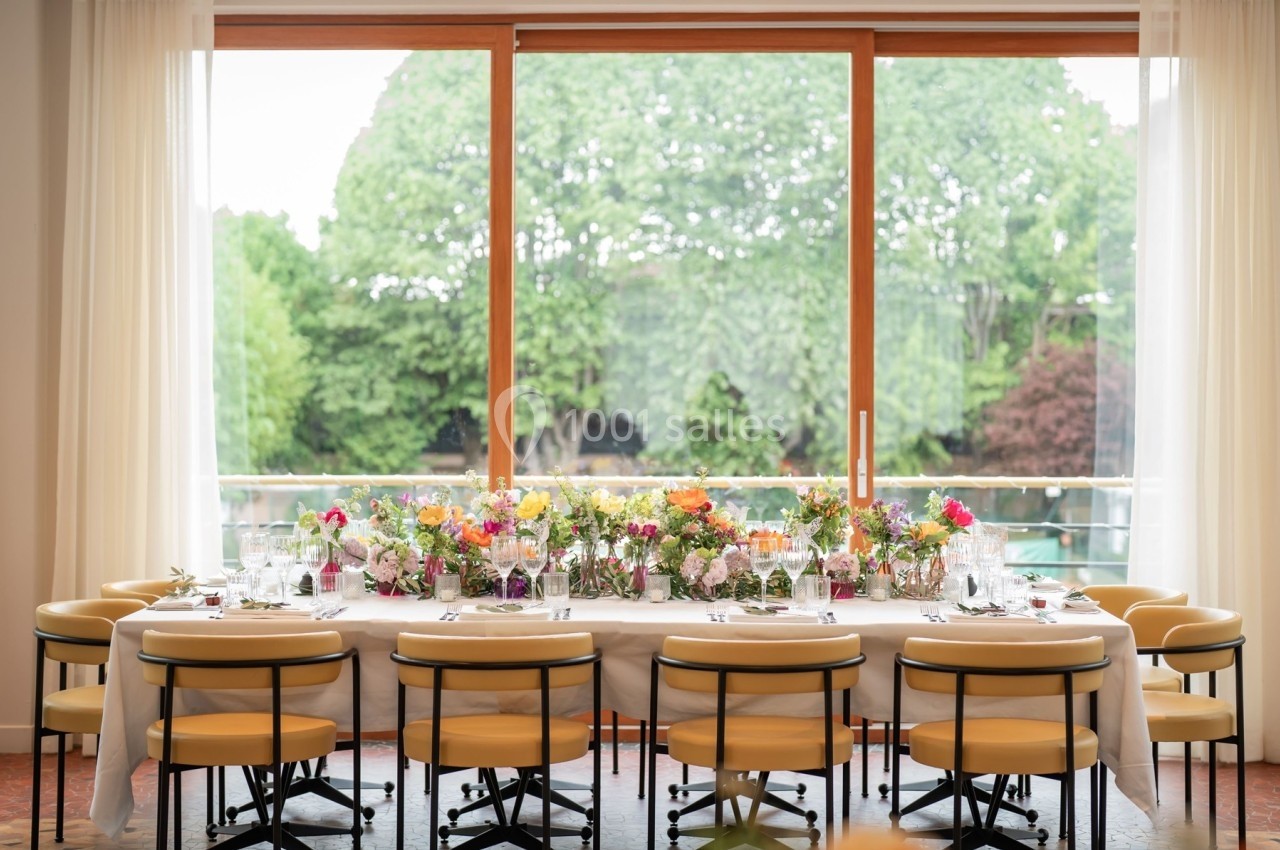 Table dressée avec des fleurs colorées, vaisselle élégante et chaises jaunes, devant une grande baie vitrée donnant sur un…