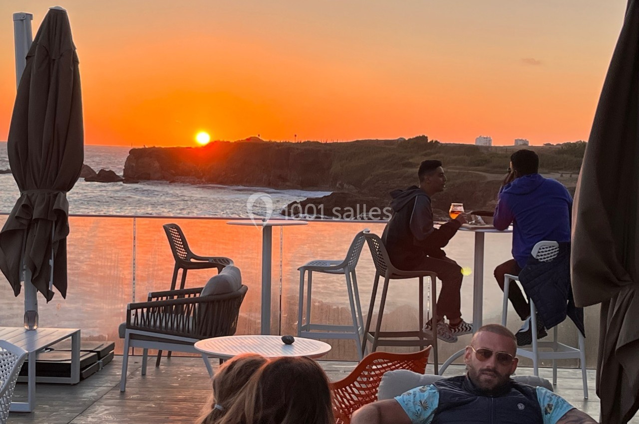 Personnes assises en terrasse face à un coucher de soleil sur la mer, avec des parasols fermés en arrière-plan.