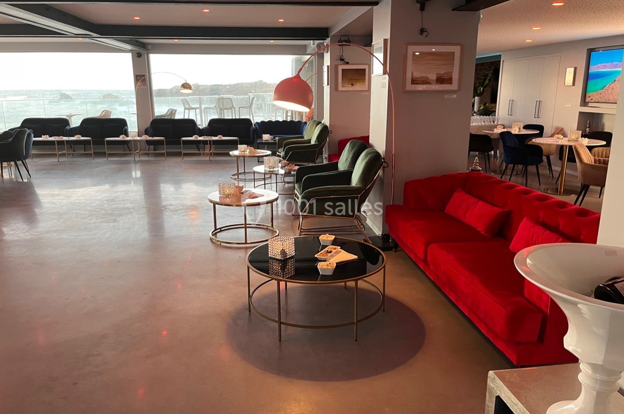 Salon moderne avec canapés rouges et verts, tables basses rondes et vue sur l'extérieur à travers de grandes baies vitrées.