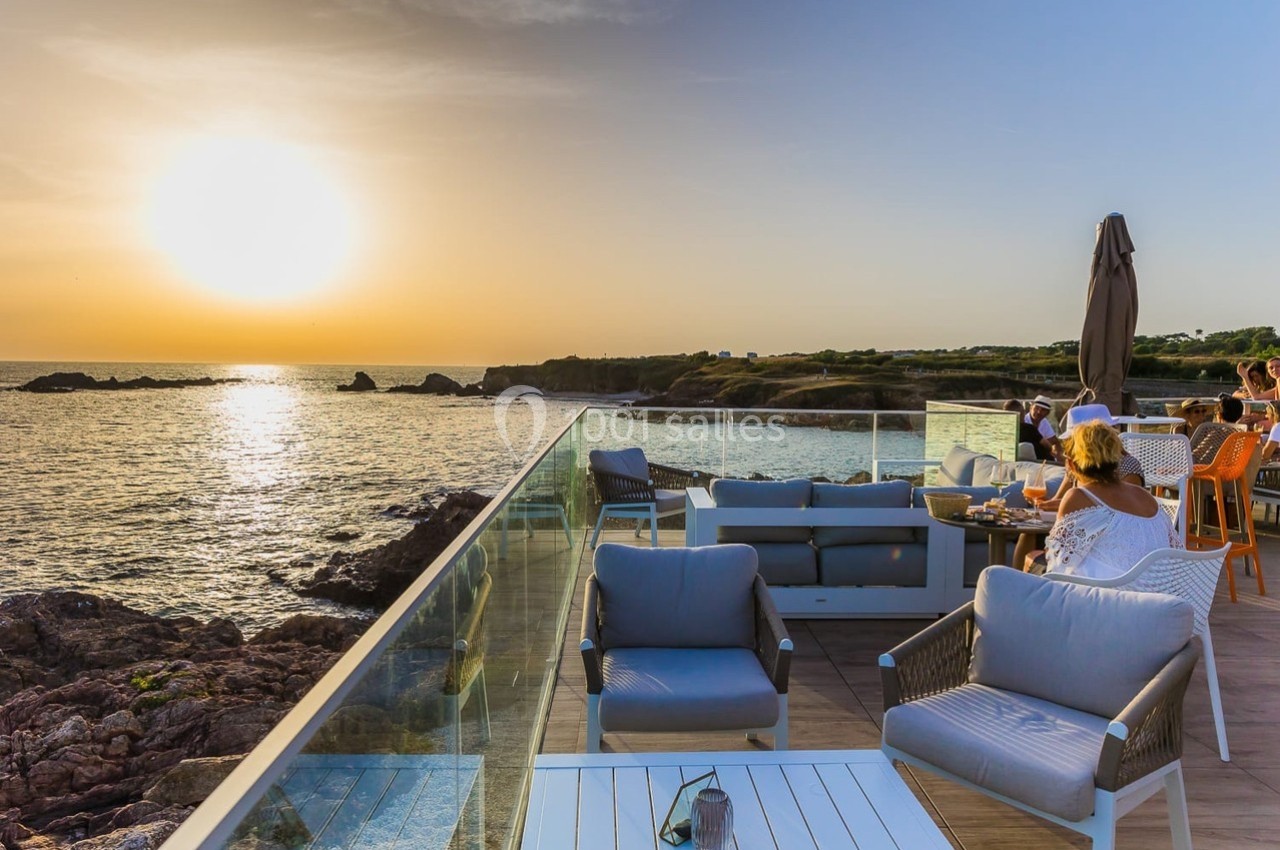 Terrasse en bord de mer au coucher du soleil, avec des tables occupées et des fauteuils face à l'horizon.
