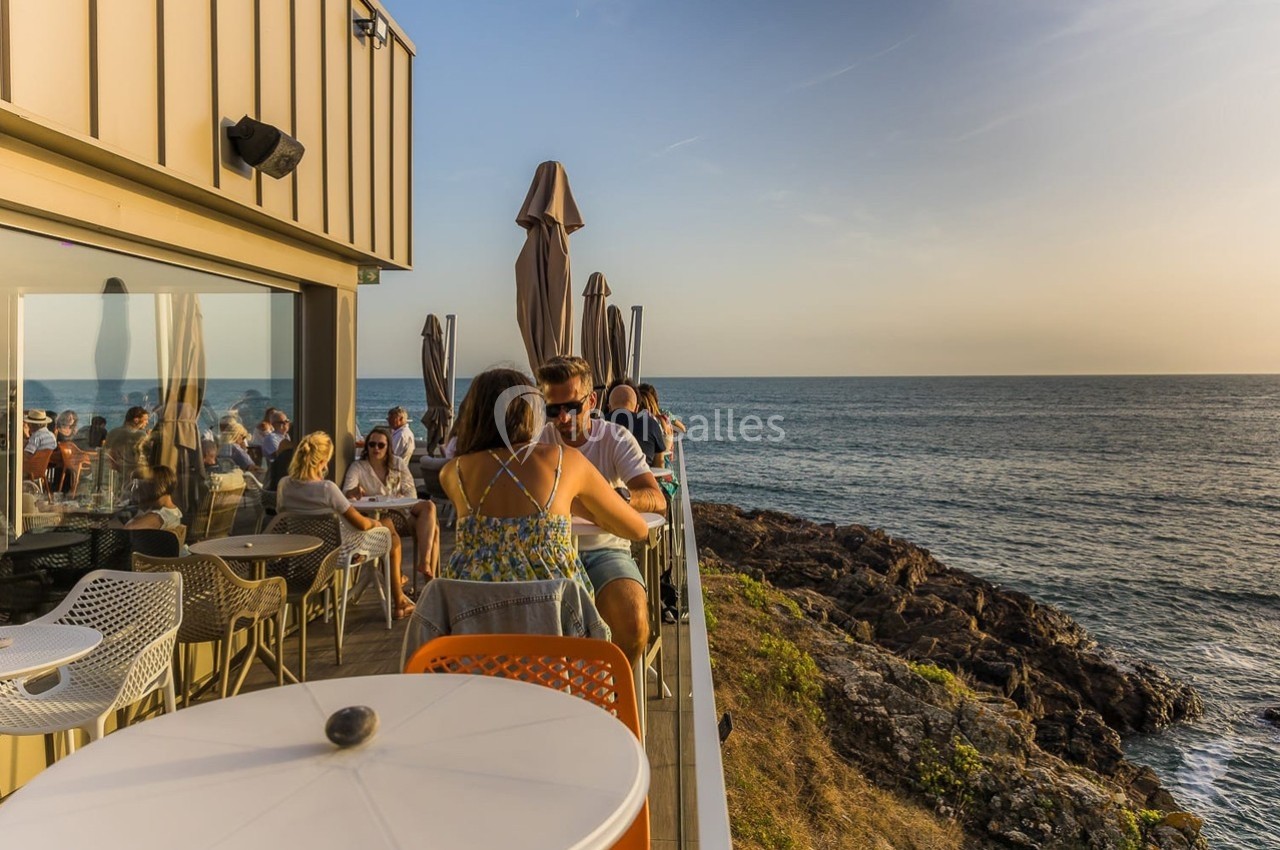 Terrasse en bord de mer avec des personnes assises, vue sur l'océan et le coucher de soleil.