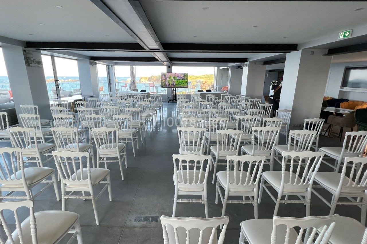 Salle lumineuse aménagée avec des rangées de chaises blanches face à un écran, vue sur la mer en arrière-plan.