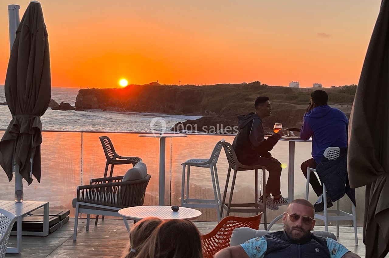 Des personnes assises en terrasse face à un coucher de soleil sur la mer, avec des parasols fermés en arrière-plan.
