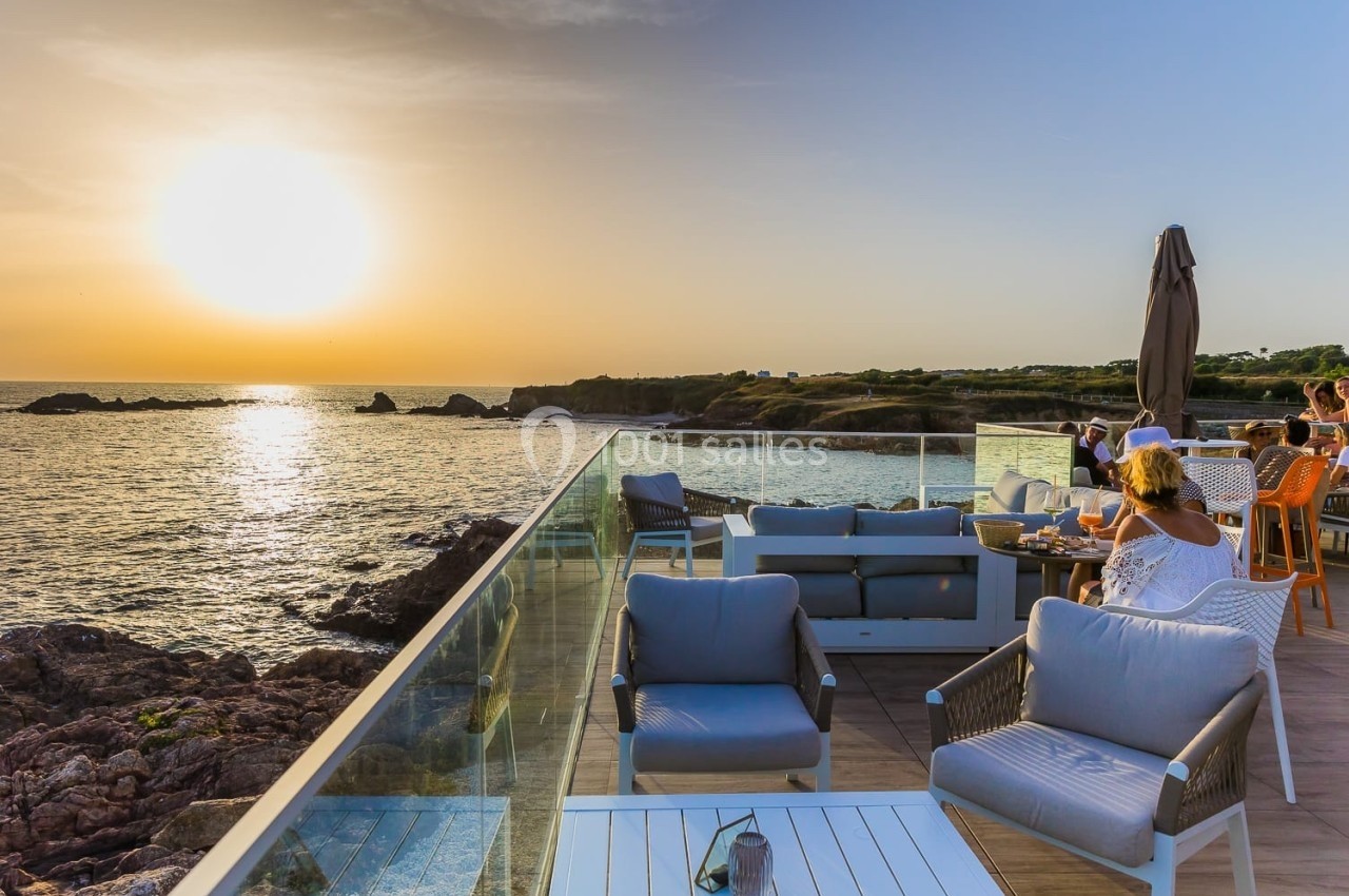 Terrasse en bord de mer au coucher du soleil, avec des tables occupées et des fauteuils vides au premier plan.