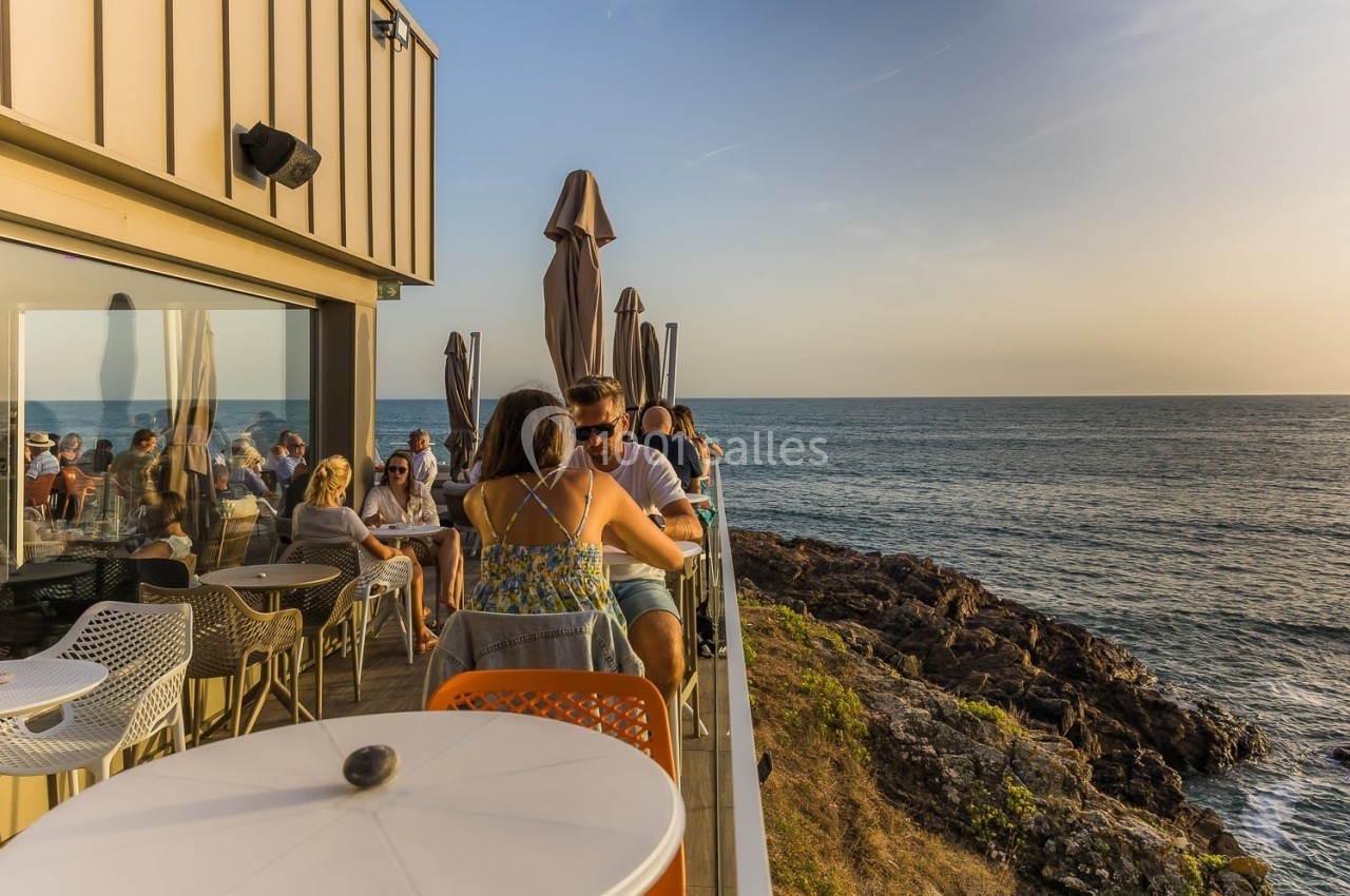 Des personnes assises en terrasse surplombant la mer au coucher du soleil, avec des parasols fermés en arrière-plan.