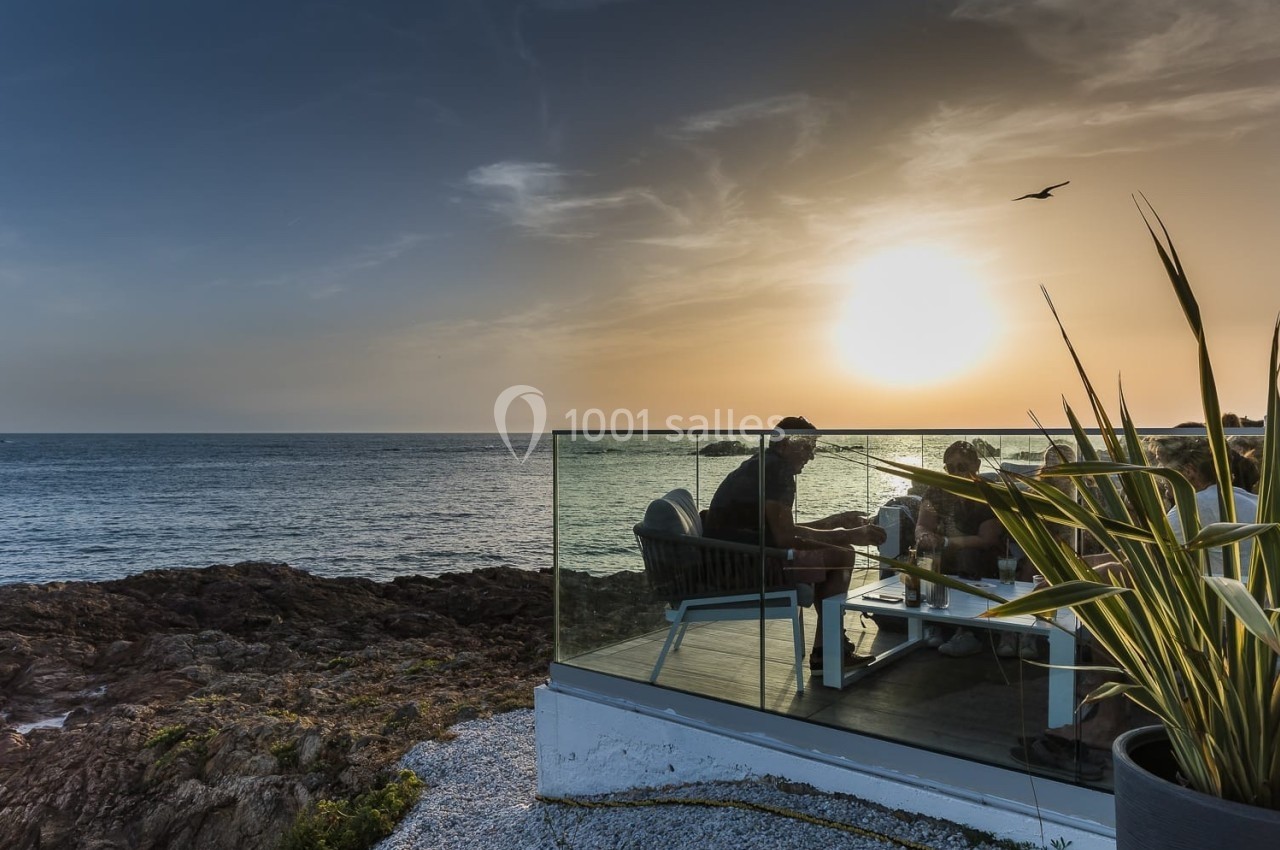 Personnes assises à une table en terrasse face à la mer au coucher du soleil, entourées de végétation et de rochers.