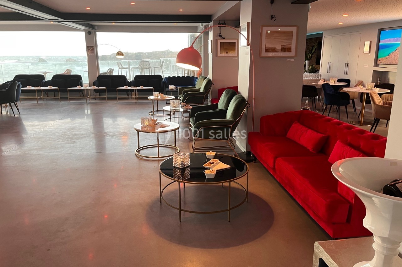 Salon moderne avec canapés rouges et verts, tables basses noires et vue sur une baie à travers de grandes baies vitrées.