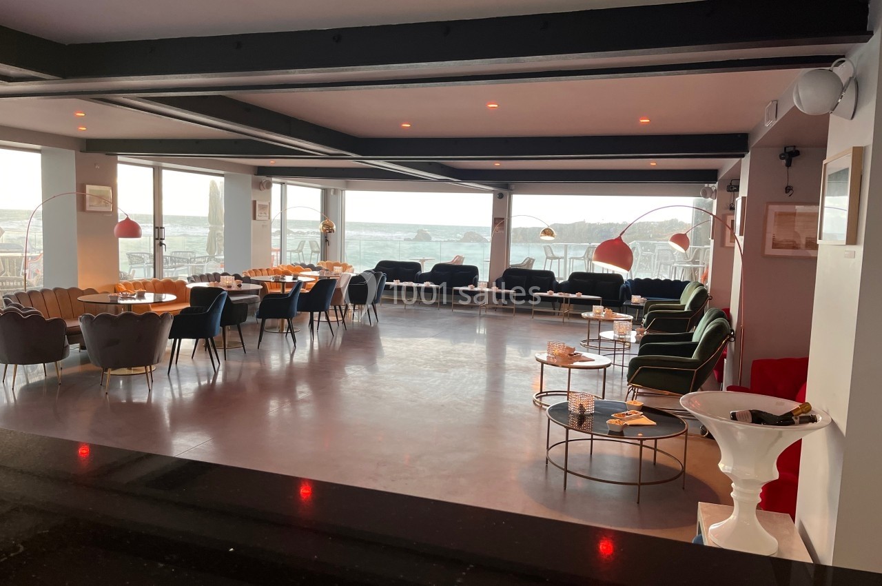 Salle lumineuse avec de grandes baies vitrées, des tables et des fauteuils disposés dans un espace moderne et épuré.
