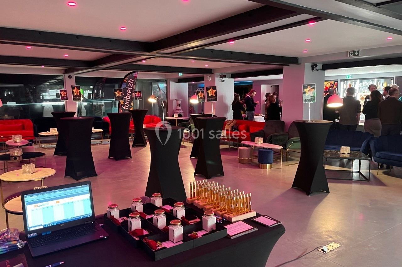 Salle moderne avec tables hautes, décor lumineux et buffet disposé, des personnes discutent en arrière-plan.
