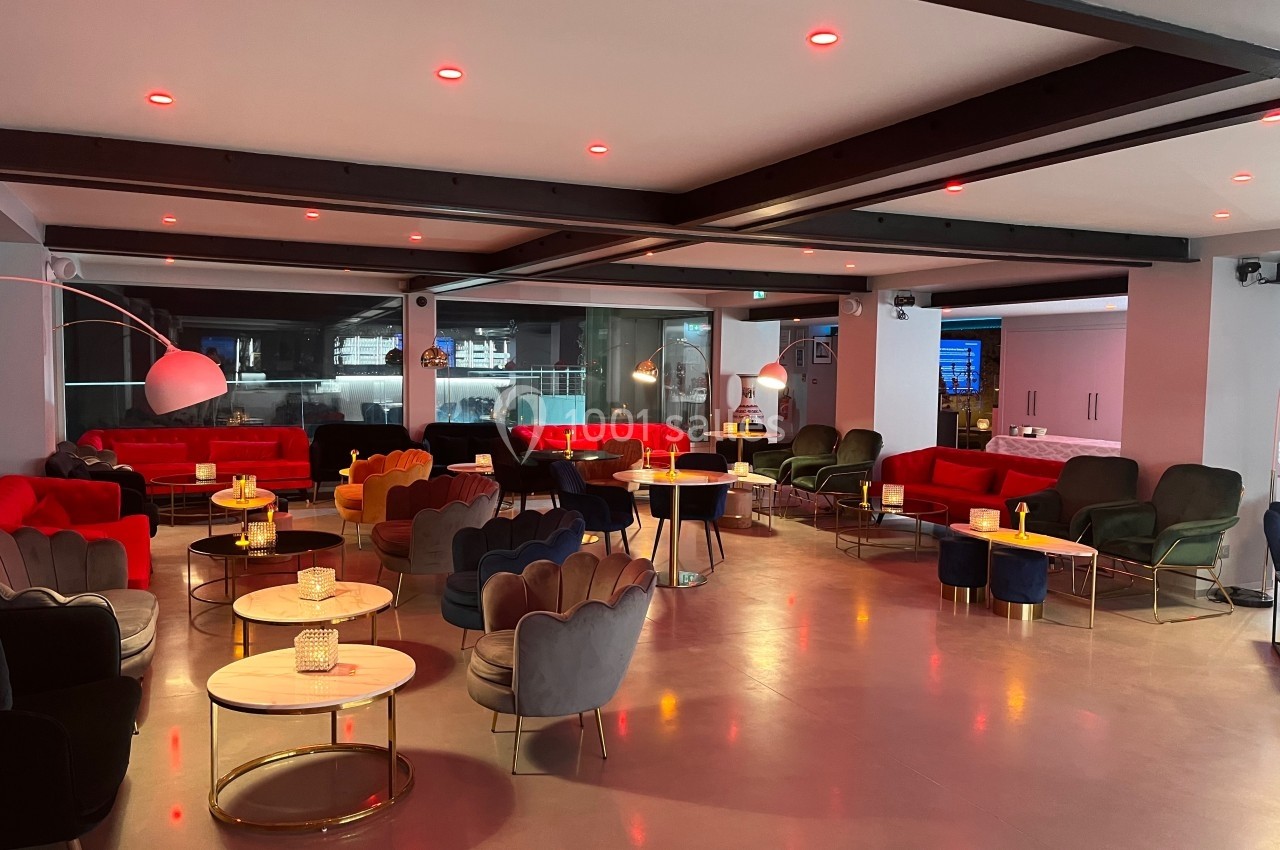 Salle moderne avec des fauteuils colorés, tables basses rondes, éclairage tamisé et grandes baies vitrées.