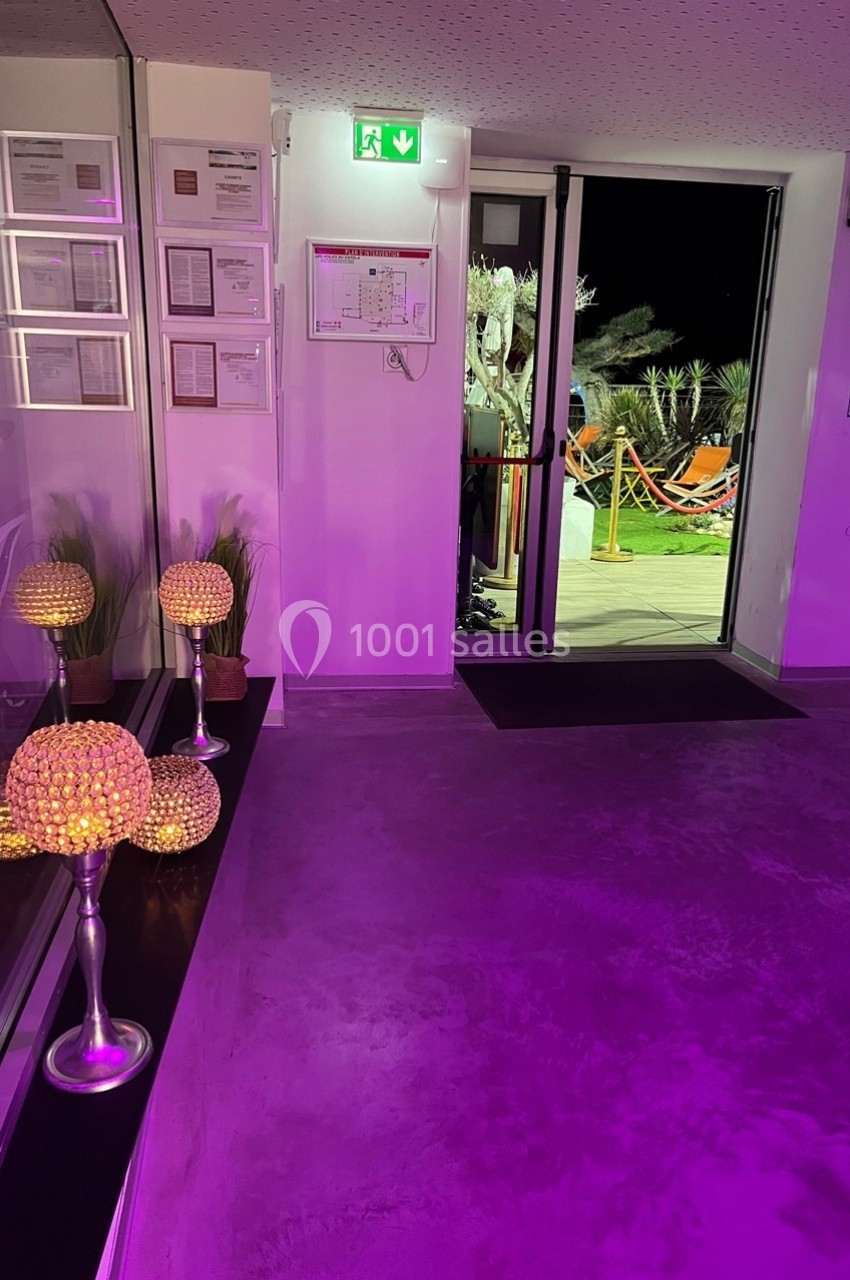 Entrée d'un bâtiment avec éclairage violet, documents encadrés au mur et porte ouverte donnant sur une terrasse éclairée.