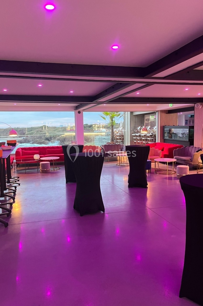 Salle moderne avec grandes baies vitrées, tables hautes noires, canapés colorés et éclairage rose tamisé.