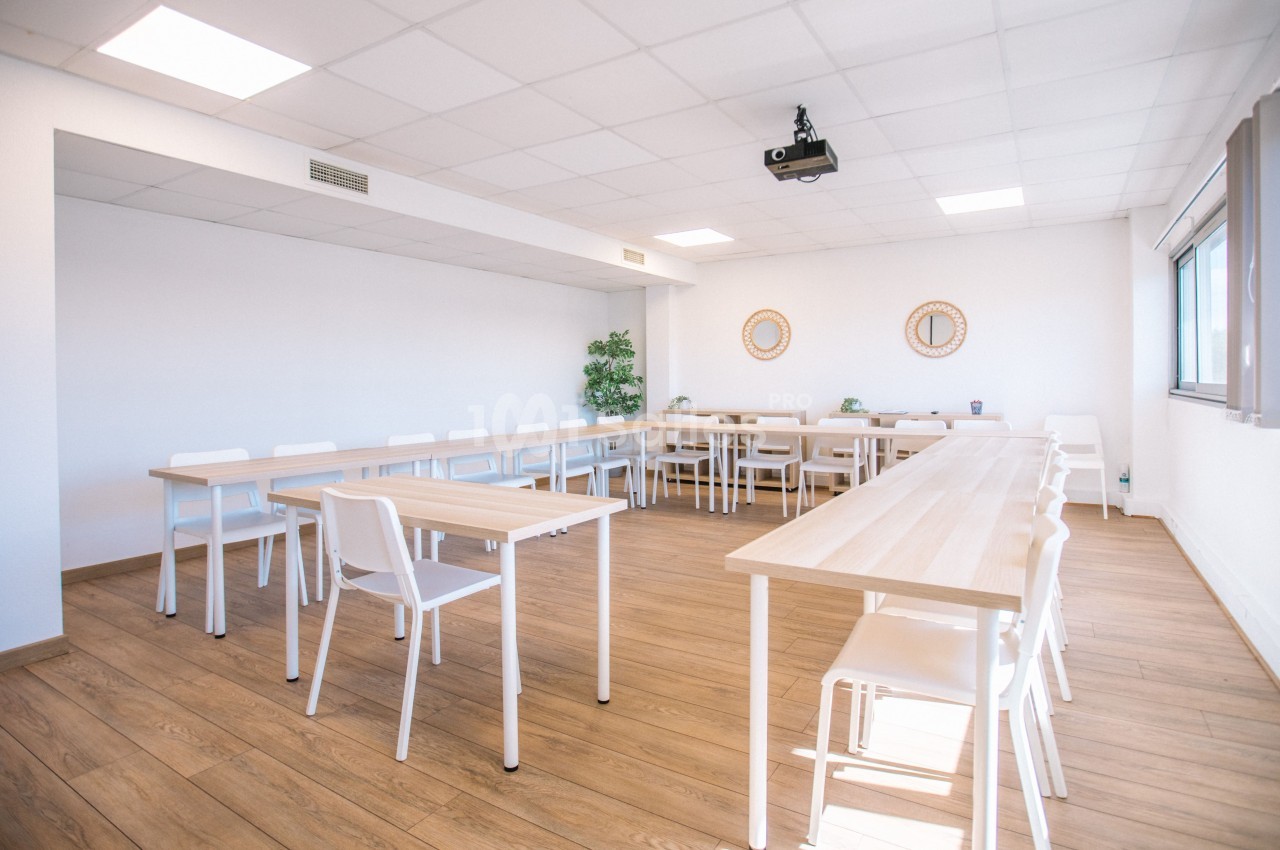 Salle lumineuse avec tables en bois clair disposées en U, chaises blanches, parquet et projecteur au plafond.