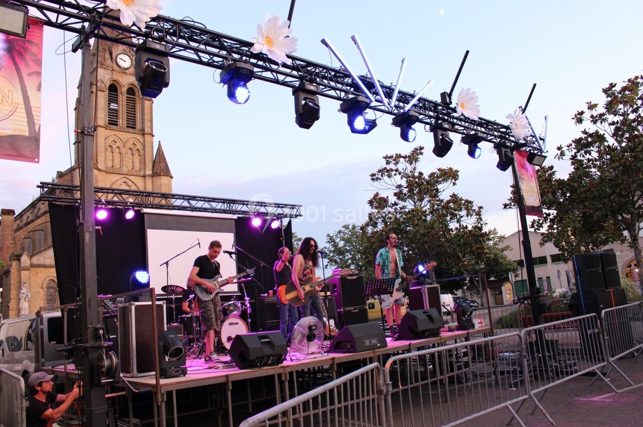 Groupe de musiciens jouant sur une scène extérieure éclairée, avec une église en arrière-plan au crépuscule.