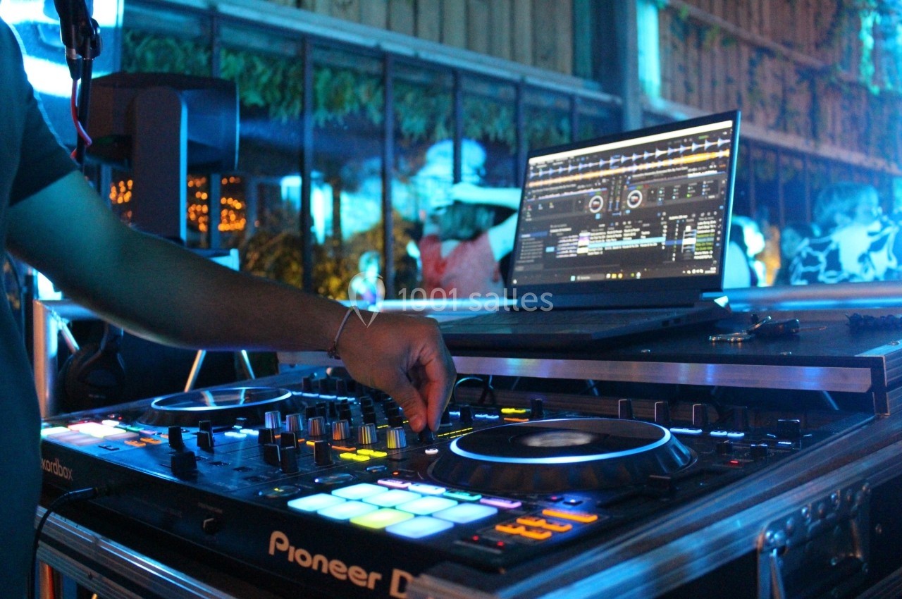 Un DJ manipule une table de mixage éclairée, avec un ordinateur affichant un logiciel de musique en arrière-plan.