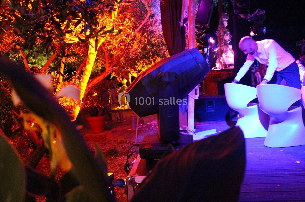Un homme ajuste des chaises blanches sur une terrasse éclairée par des lumières colorées, entourée de végétation.