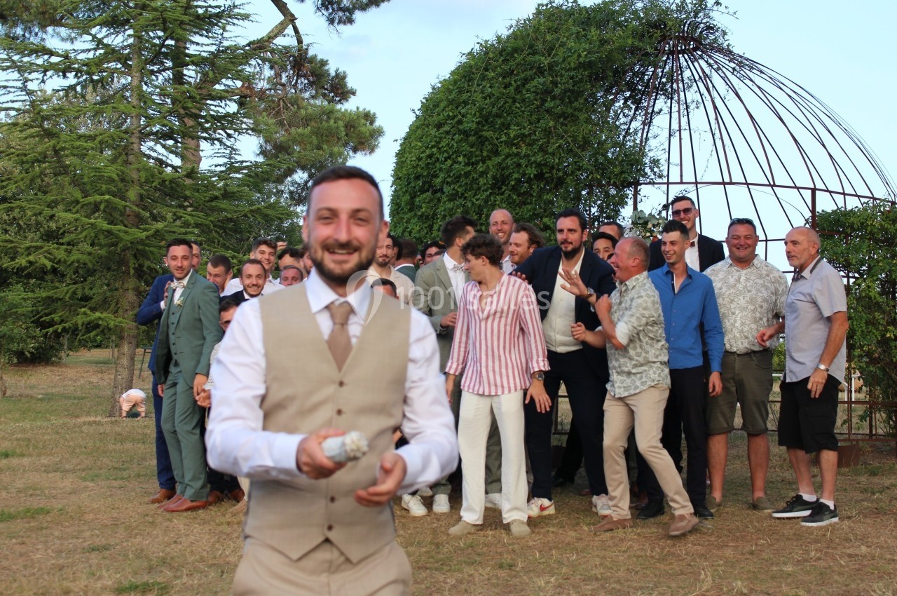 Un homme en costume beige sourit en tenant un objet, avec un groupe d'hommes en arrière-plan dans un jardin.
