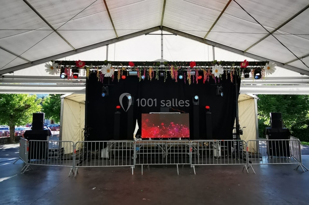 Scène couverte en plein air avec équipement sonore, décorations florales suspendues et barrières de sécurité.