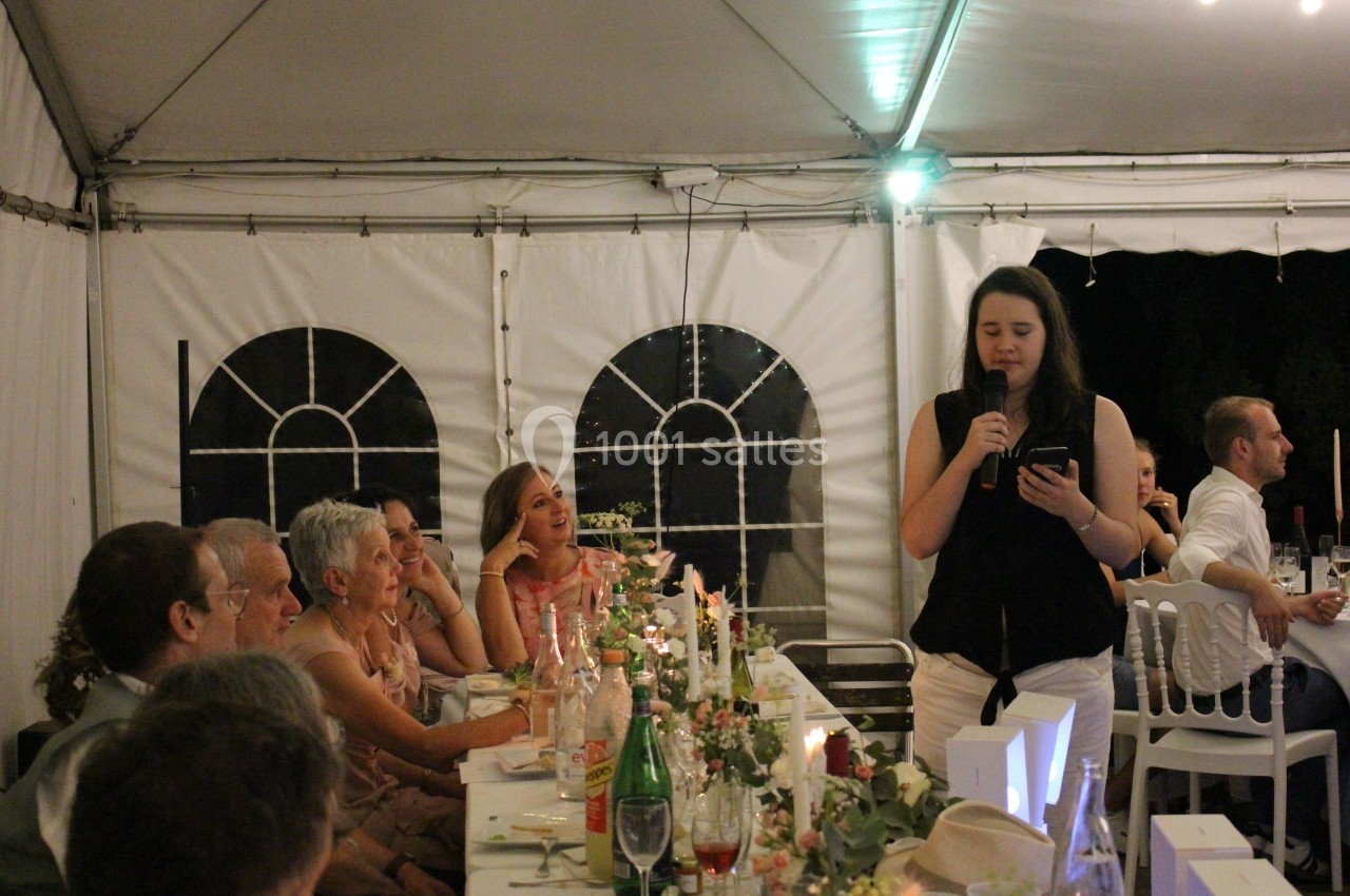 Une femme fait un discours au micro lors d'un dîner sous une tente, entourée de convives attentifs.