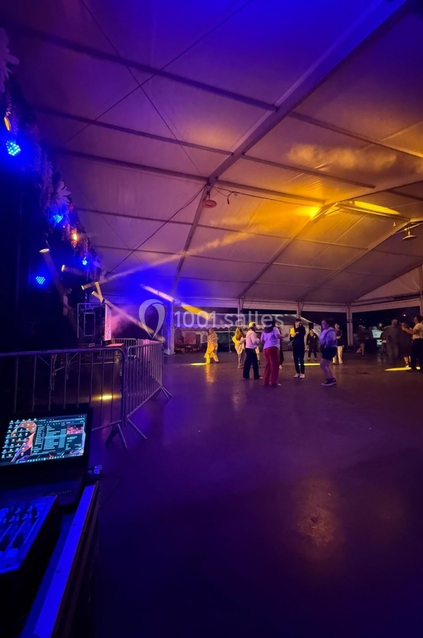 Des personnes dansent sous un chapiteau éclairé par des lumières colorées, avec une table de mixage au premier plan.