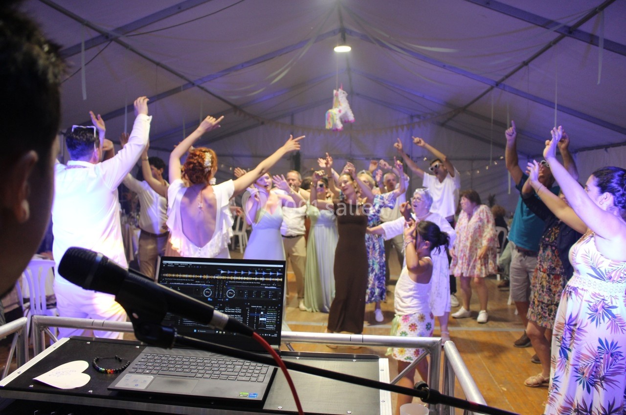 Groupe de personnes dansant sous une tente éclairée, vu depuis une table de mixage avec un micro au premier plan.