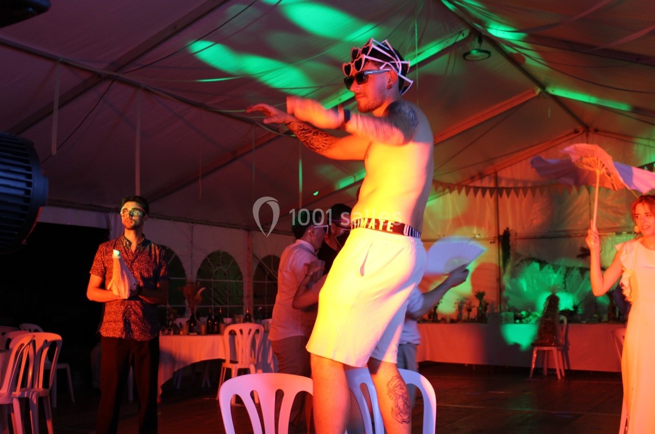 Un homme en tenue décontractée danse sur une table dans une tente éclairée par des lumières colorées, entouré de spectateurs.