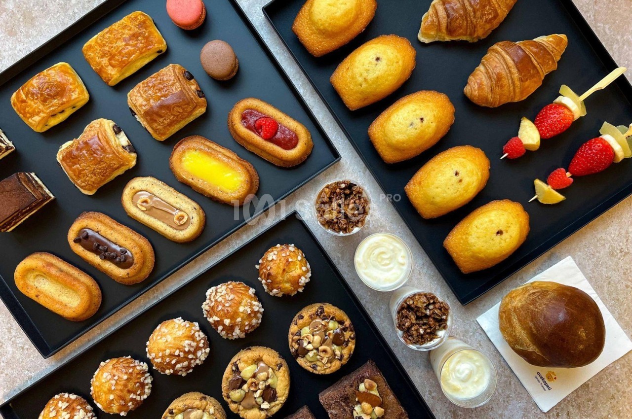 Assortiment de viennoiseries, pâtisseries, muffins, madeleines et desserts présentés sur des plateaux noirs.