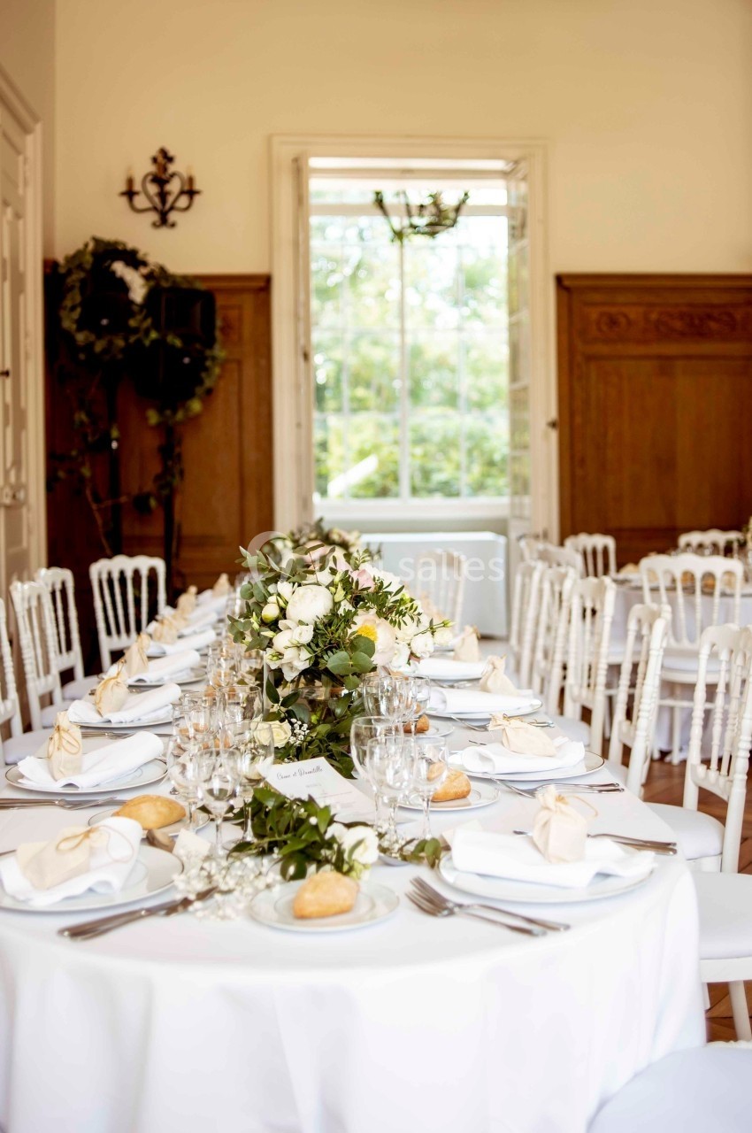 Salle de réception lumineuse avec tables rondes dressées, décorées de fleurs blanches et verdure.