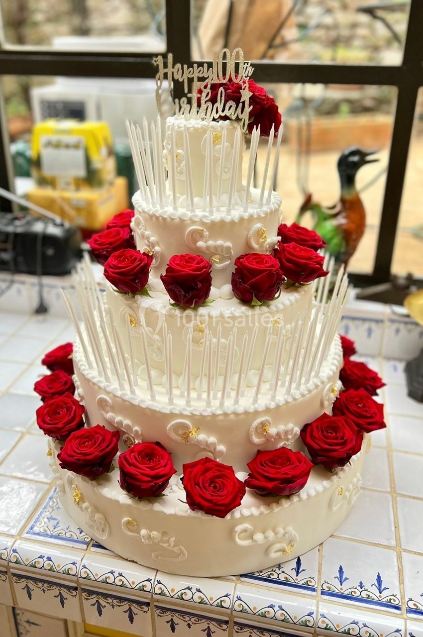 Gâteau de fête à trois étages décoré de roses rouges et de détails blancs, avec une inscription ’Happy 100th Birthday’.