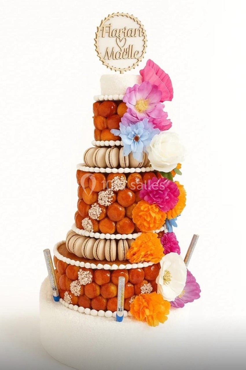 Pièce montée composée de choux, macarons et fleurs colorées, ornée d’un disque personnalisé au sommet.