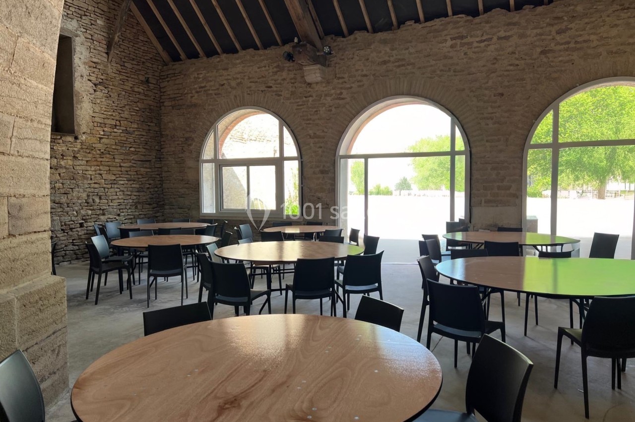Salle avec des murs en pierre, des tables rondes en bois et des chaises noires, éclairée par de grandes fenêtres cintrées.