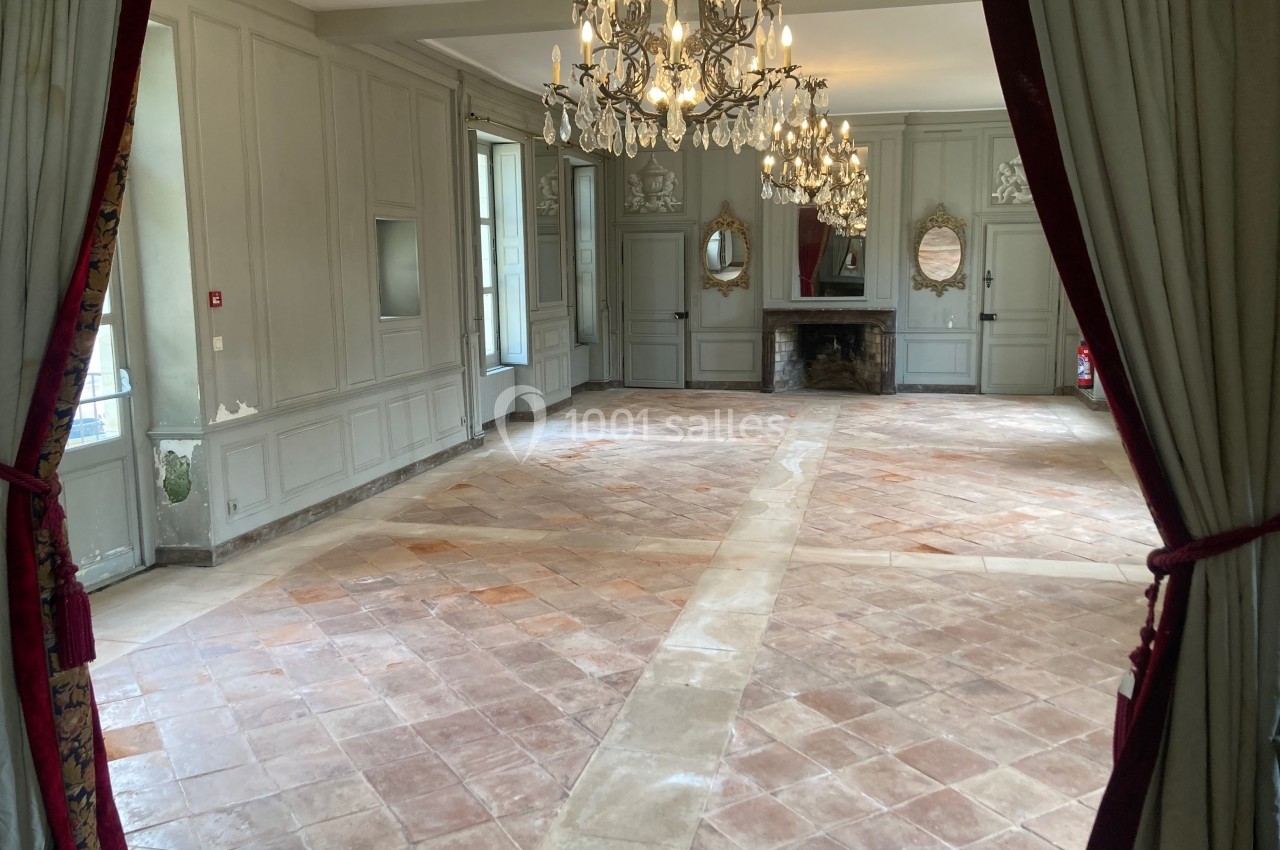 Salle vide avec sol en carreaux de terre cuite, murs clairs, cheminée, miroirs et lustres suspendus.