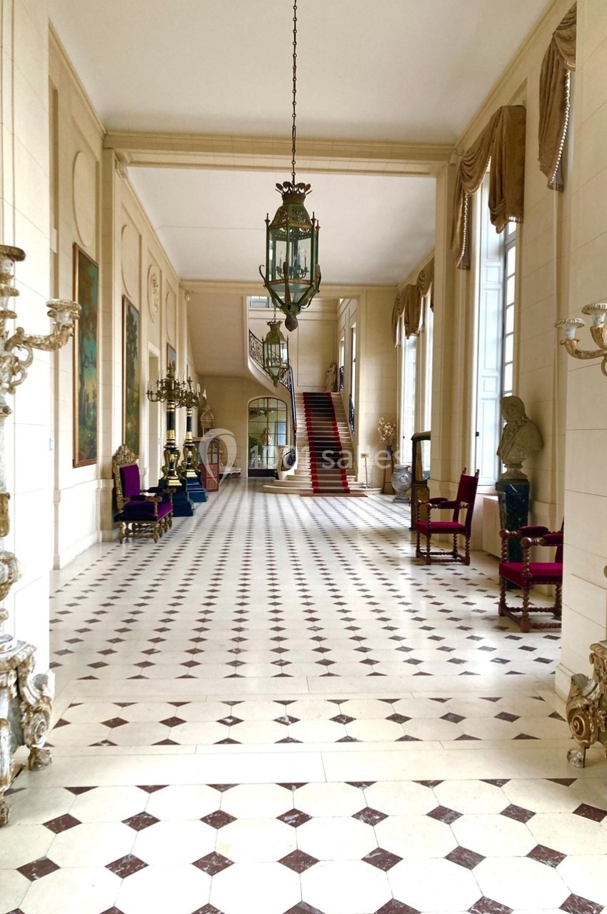 Hall spacieux avec sol en damier, lustres anciens, chaises décoratives et escalier au fond.