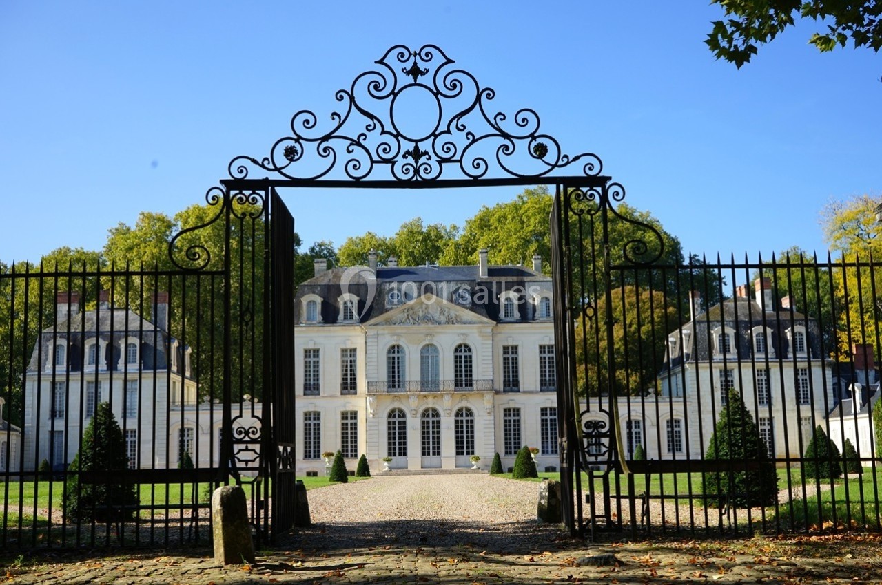 Grille en fer forgé ouvrant sur un château blanc entouré d'arbres sous un ciel bleu.