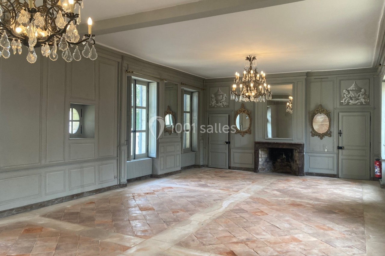 Salle lumineuse avec murs gris ornés de miroirs, cheminée en pierre, lustres en cristal et sol en carreaux de terre cuite.