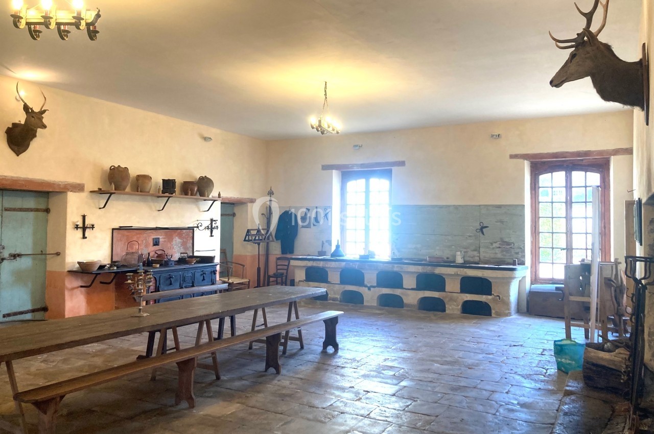 Cuisine rustique avec longue table en bois, fourneau ancien, étagères en terre cuite et murs ornés de têtes de cerfs.