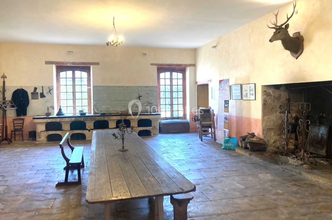 Grande cuisine ancienne avec une longue table en bois, cheminée, cuisinières en pierre et décoration murale.