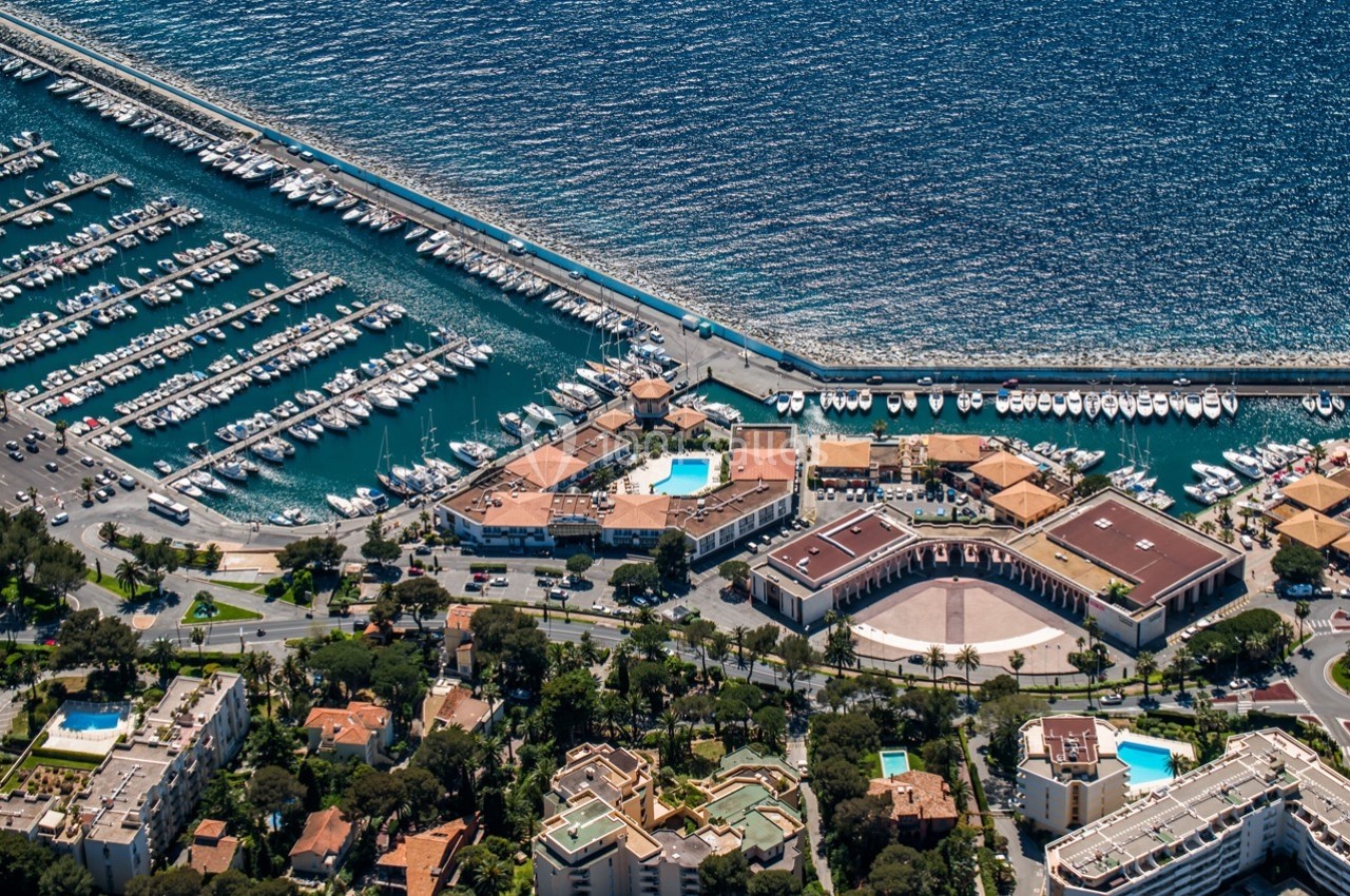 Vue aérienne d'un port de plaisance avec des bateaux amarrés, entouré de bâtiments et d'une route côtière.